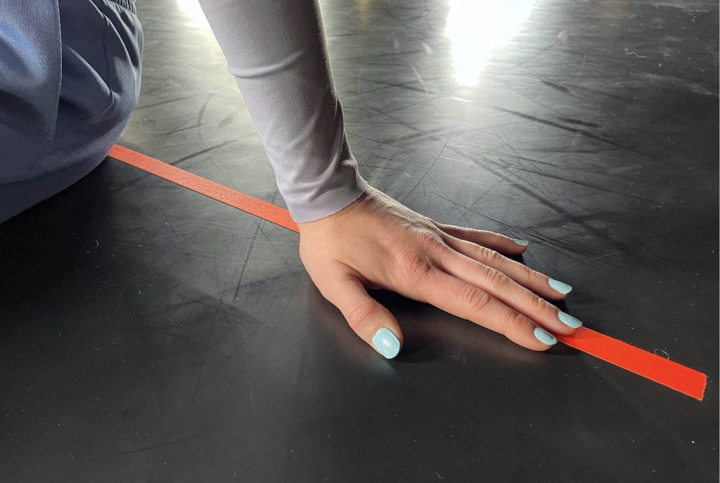 A close up image of a hand with blue coloured nails resting on the floor marked with an orange tape. 